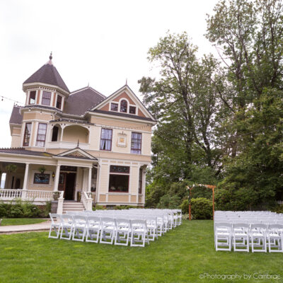 A wedding ceremony is set on a lawn in front of green trees and an ornate Victorian mansion.