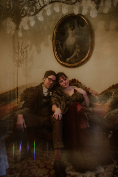 A man and a woman sit on an antique couch under a vintage painting of a woman