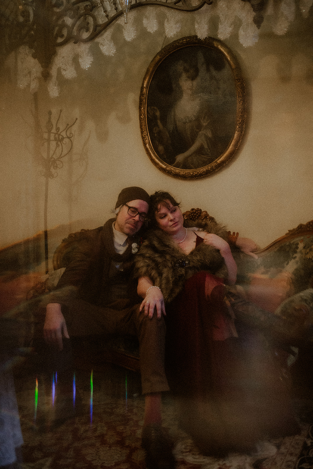 A man and a woman sit on an antique couch under a vintage painting of a woman