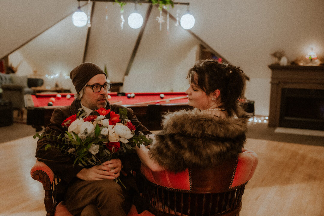 A man and a woman sit facing each other in a tete-a-tete chair. The man holds a bouquet of flowers