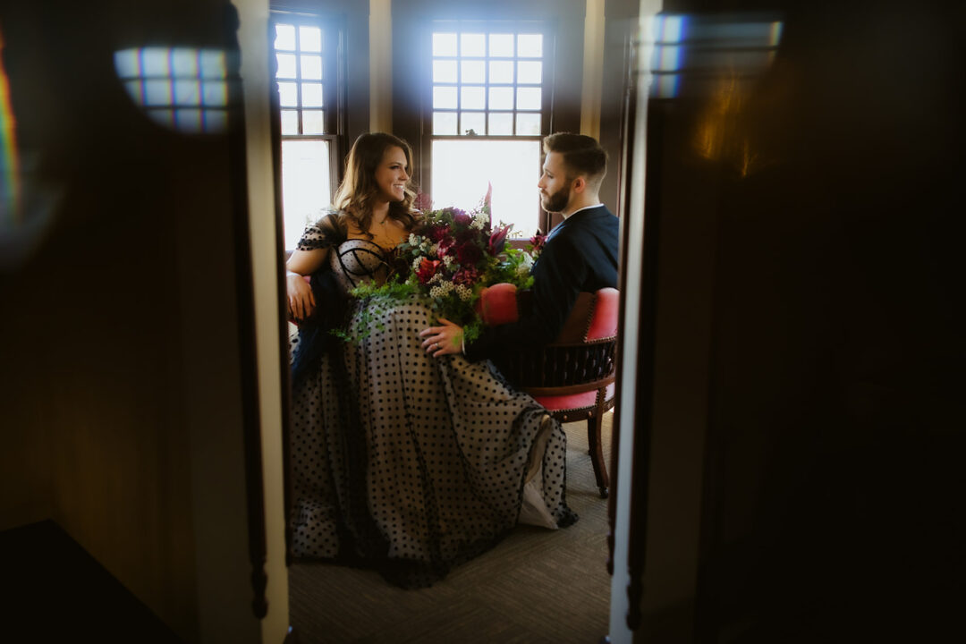 A bride and groom face each other on a vintage pink chair in the turret of the Victorian Belle Mansion in North Portland. The bride wears a black lace dress and the groom wears a black suit. Photo by Megan Gallagher