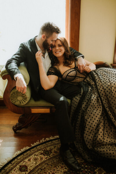A bride and groom lounge together on a velvet green sofa at the Victorian Belle Mansion in Portland, Oregon. The bride wears a black lace gown and the groom wears a black suit. Photo by Megan Gallagher.