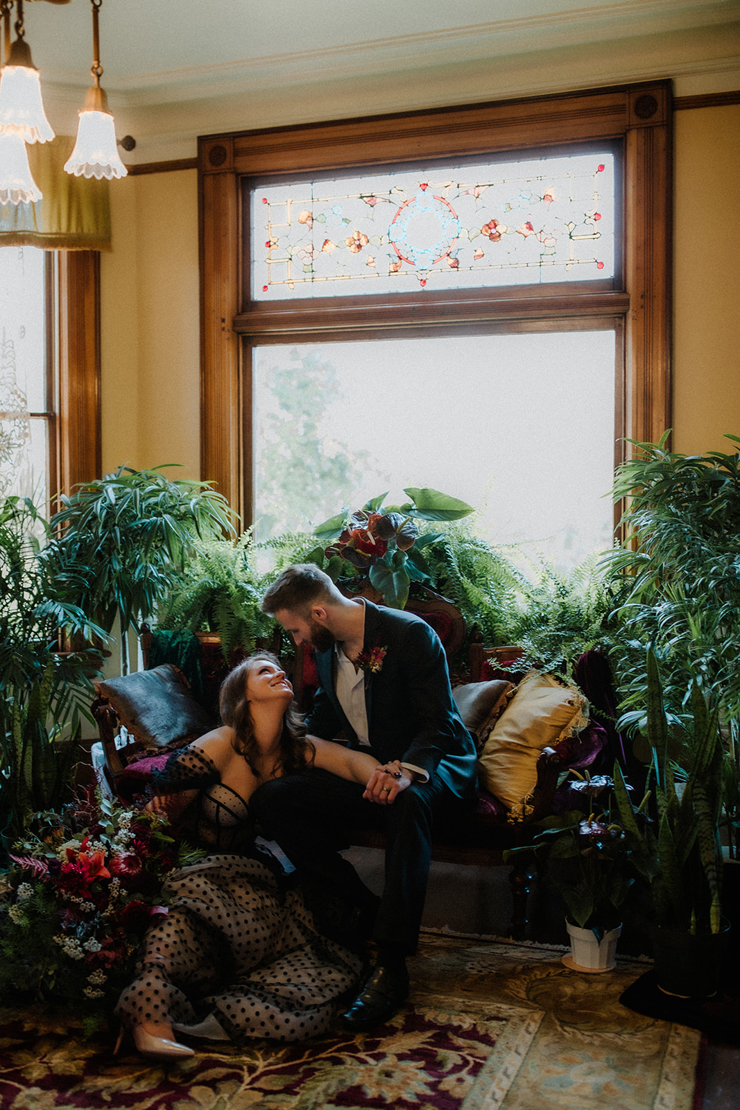 A bride and a groom sit in the parlor of the Victorian Belle Mansion surrounded by tall leafy potted plants and stained glass windows. Photo by Megan Gallagher