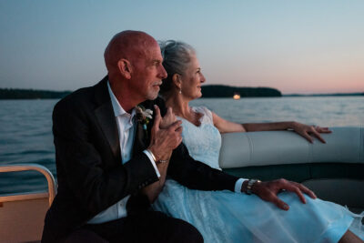 A mature couple sits on the white leather seats of a pontoon boat on a lake in the sunset. The fading light casts a warm glow on their faces--the groom has a bald head and is wearing a black tux. The bride has long silver hair twisted into an up-do, with strands blowing in the wind; her dress is a light blue, retro lace style. They are leaning into each other as they look to the sunset, holding hands. It very much looks like a movie poster!