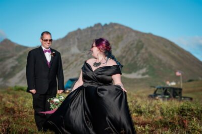 A bride in a striking black satin dress is laughing and smiling and looking back to her partner, a groom in a black tux, as they are moving across an alpine meadow in Alaska. The brides hair is purple/rainbow, and the groom's bowtie and vest match her hair! All around them, ground-level, flat bushes, tinted red in fall colors, are filled with wild blueberries at their feet. Behind them large mountains rise, and you can see the tops of their UTV machines they drove up the mountain peeking over the mounds of the foliage behind them.