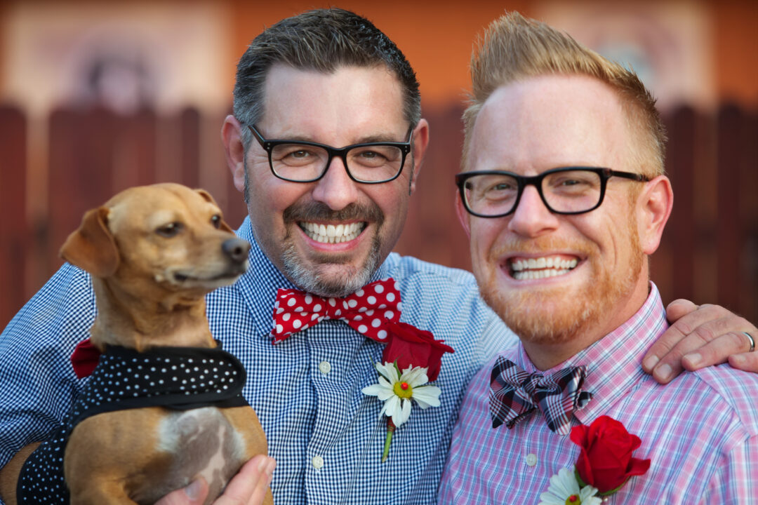Two smiling grooms wearing glasses and colorful bow ties pose outdoors with their small brown dog, who is dressed in a polka-dot bandana.