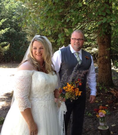 Bride and groom stand together outdoors holding a bouquet during a rustic wedding ceremony.