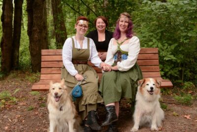 Couple in Renaissance-inspired attire sit on a bench with their two dogs while their officiant stands smiling behind them in a forest setting.