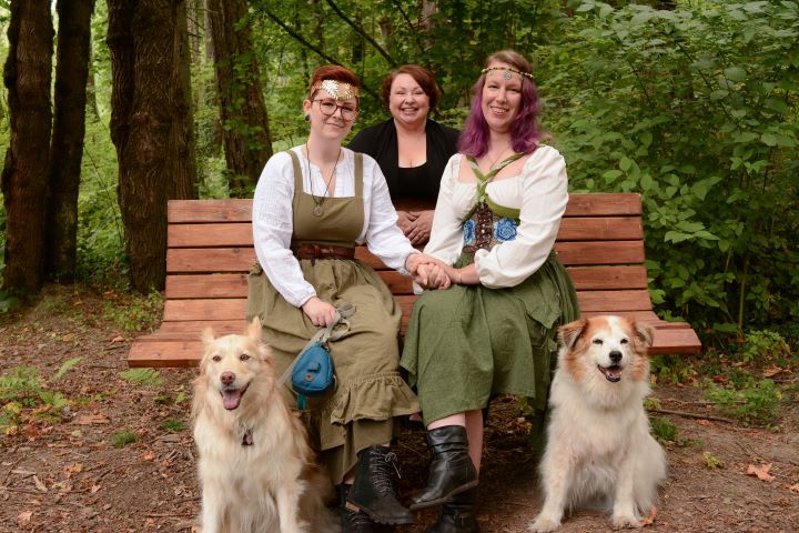 Couple in Renaissance-inspired attire sit on a bench with their two dogs while their officiant stands smiling behind them in a forest setting.