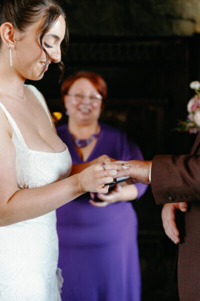 Bride places a wedding ring on her partner’s finger during an indoor ceremony as the officiant smiles in the background.