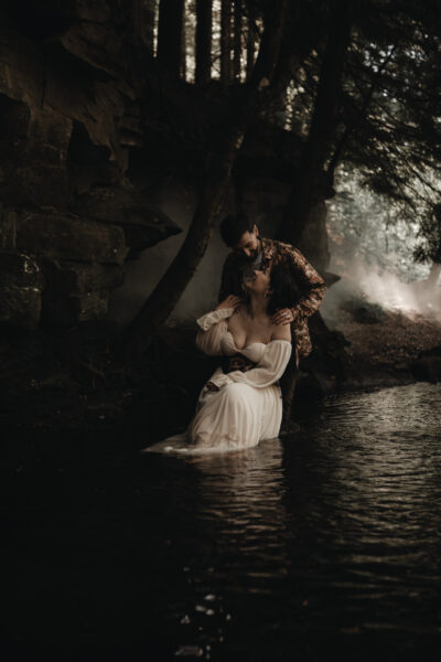 Couple embracing in a shallow forest stream beneath dark rock formations, surrounded by mist and trees, with the woman in a flowing off-shoulder white dress and the man standing behind her, creating a moody, cinematic, intimate scene.