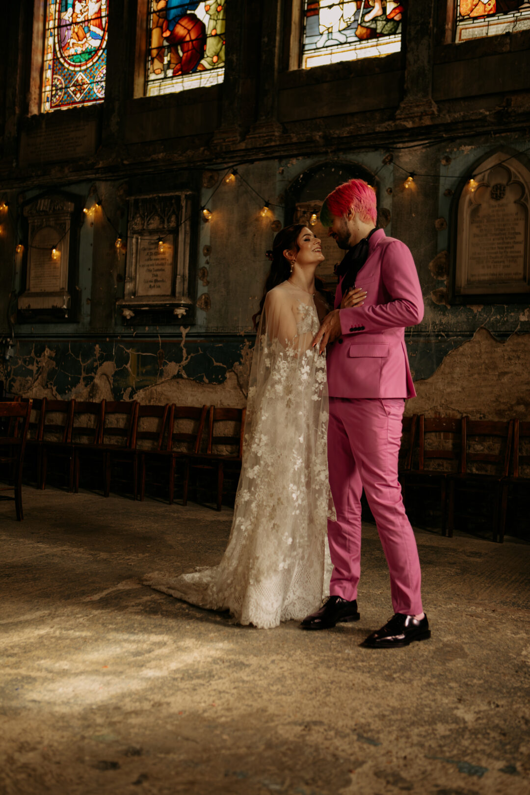 Couple standing together inside a characterful, historic church with stained glass windows and worn walls, smiling at each other, with the bride in a lace gown and the groom in a bold pink suit, creating a warm, alternative and intimate moment.
