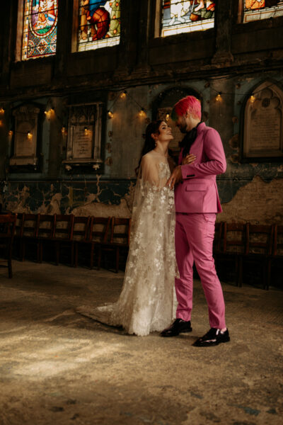 Couple standing together inside a characterful, historic church with stained glass windows and worn walls, smiling at each other, with the bride in a lace gown and the groom in a bold pink suit, creating a warm, alternative and intimate moment.