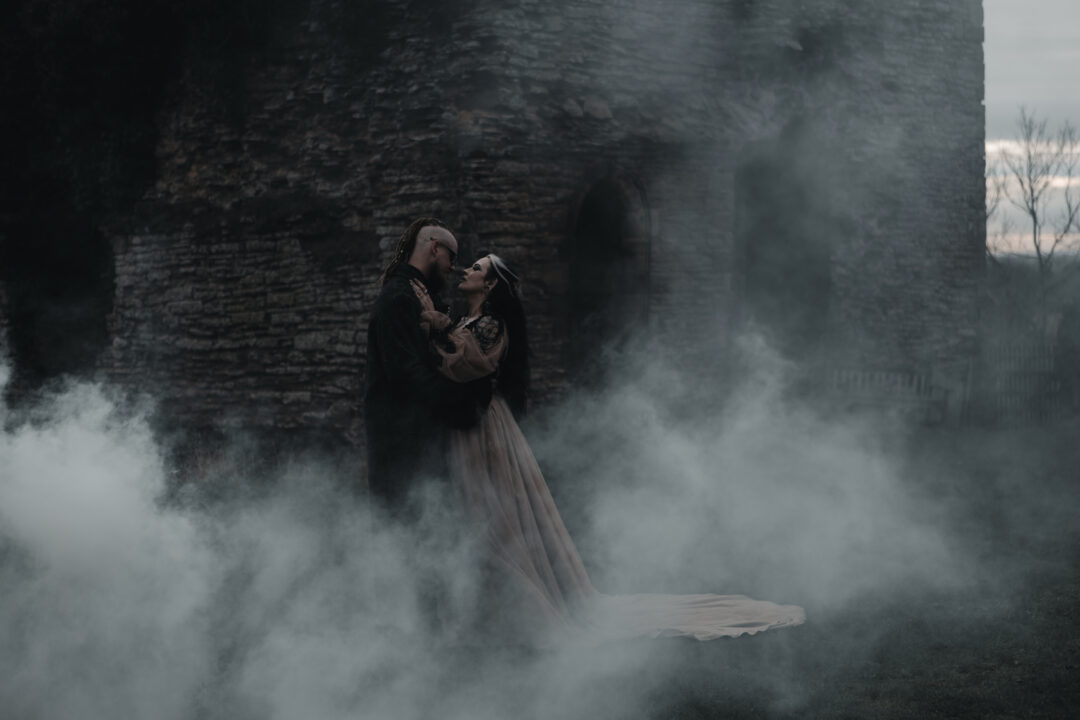 Couple standing closely together in front of an old stone tower, surrounded by drifting mist, dressed in dark and flowing attire, sharing an intimate moment in a moody, gothic setting.