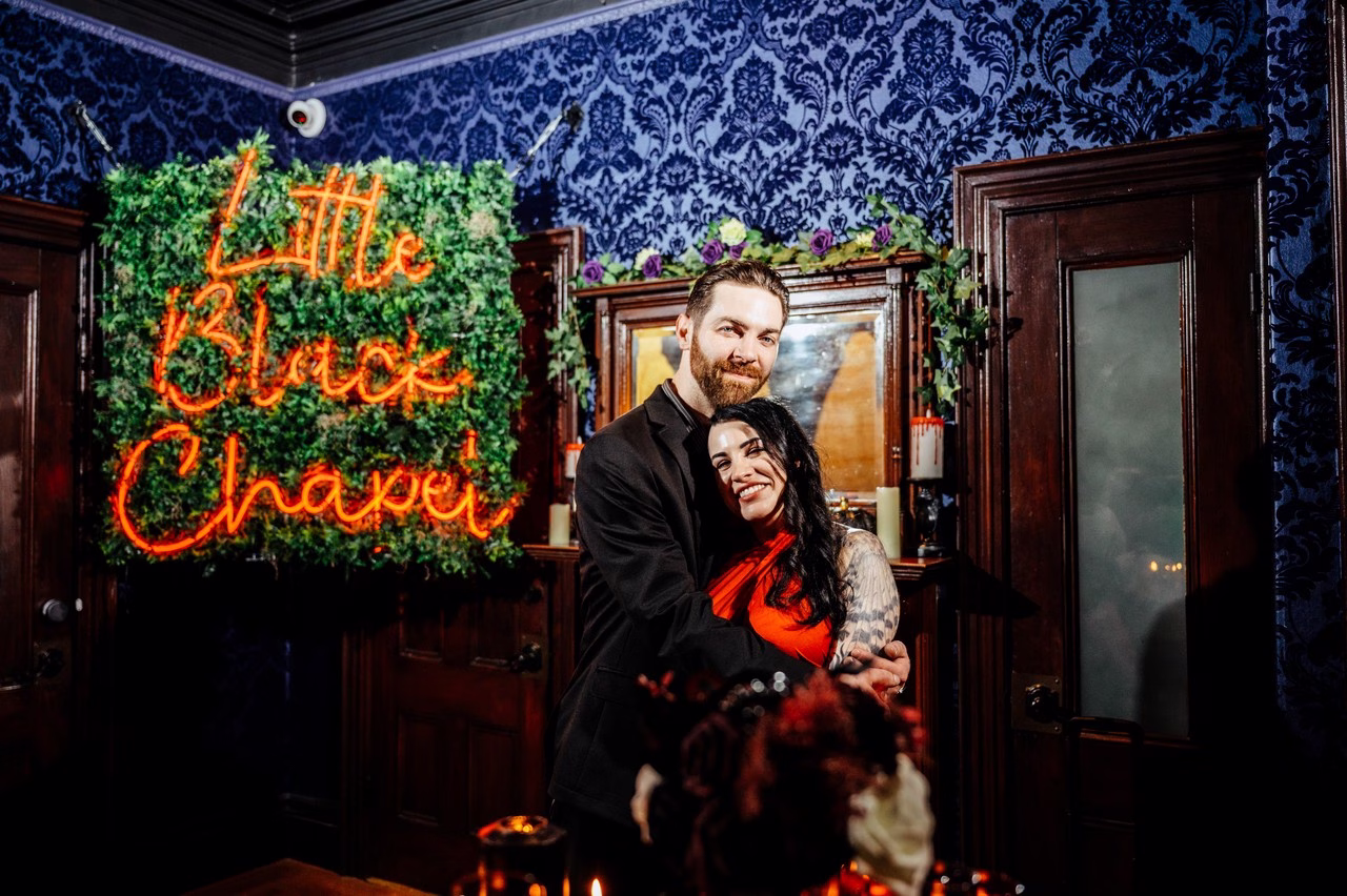 A couple embraces and smiles at the Little Black Chapel, a gothic wedding venue with blue damask wallpaper, neon signage, and candlelit decor; one wears a red dress with tattoos, the other a black suit.