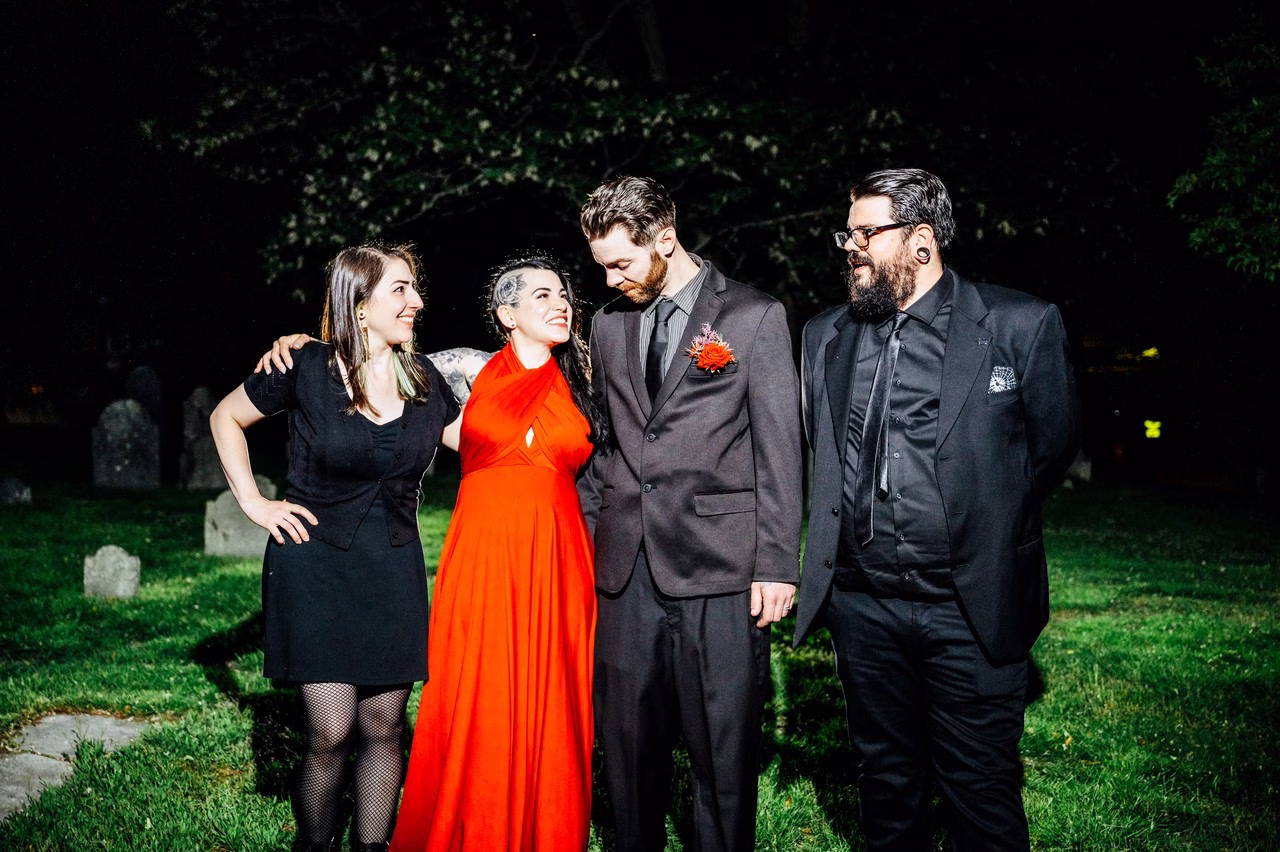 Two wedding guests in black stand with a bride in a red halter gown and groom in a gray suit with boutonniere, posing together at night in a cemetery at The Satanic Chapel, photo by Katlyn Reilly Photography