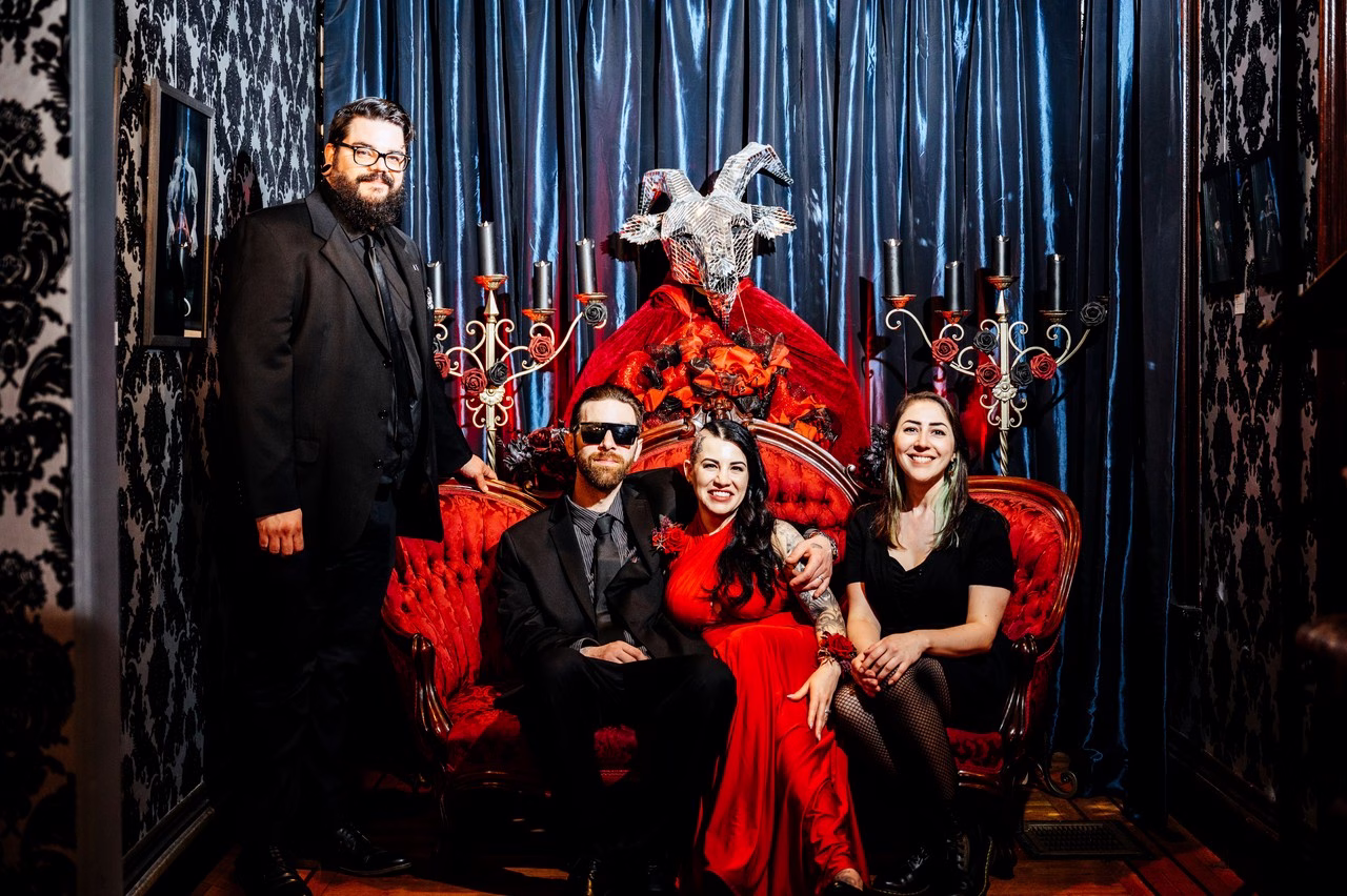 Wedding party posing in front of a dramatic red velvet throne with a silver horned figure, dark damask wallpaper, and blue satin curtains at The Satanic Chapel, photographed by Katlyn Reilly Photography