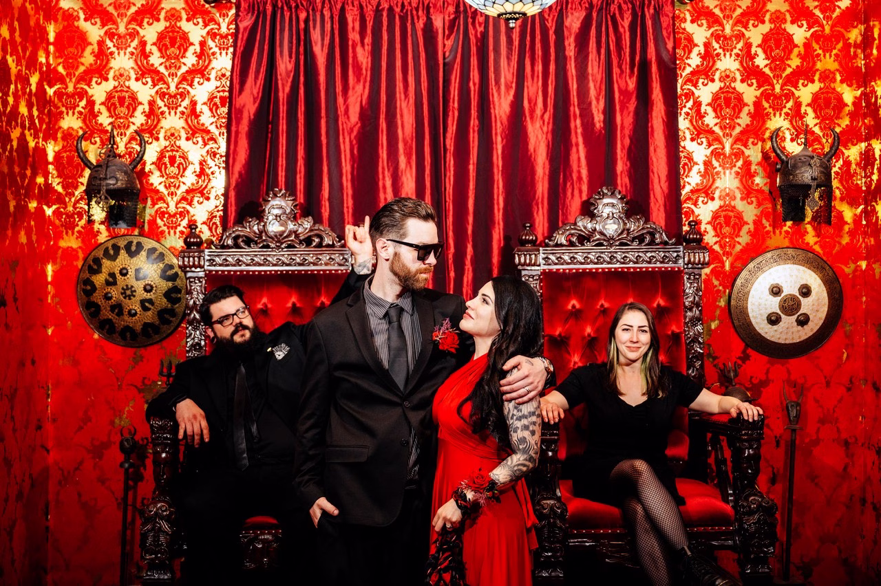 A bride in a red dress and groom in a black suit embrace in front of ornate thrones at The Satanic Chapel, with two guests seated behind them, surrounded by red damask wallpaper and decorative shields, photographed by Katlyn Reilly Photography