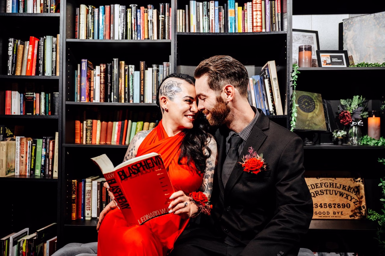 A tattooed bride in a red halter dress and groom in a black suit sit together holding a book titled Blasphemy in front of dark bookshelves with an Ouija board at The Satanic Chapel, photographed by Katlyn Reilly Photography