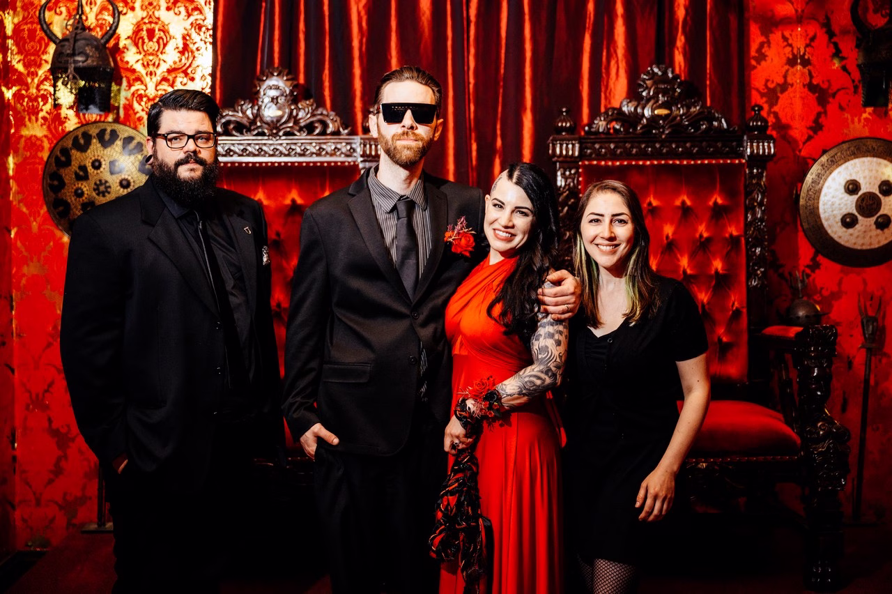 Wedding party posing in front of red velvet throne and damask decor at The Satanic Chapel, featuring bride in red gown with tattoos and groom in black suit with sunglasses, photographed by Katlyn Reilly Photography