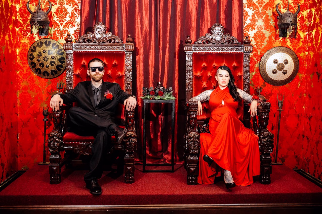 A couple sits on ornate carved wooden thrones at The Satanic Chapel, one wearing a black suit with red boutonniere and sunglasses, the other in a flowing red gown with sleeve tattoos, surrounded by red damask walls, Viking helmets, and decorative shields.