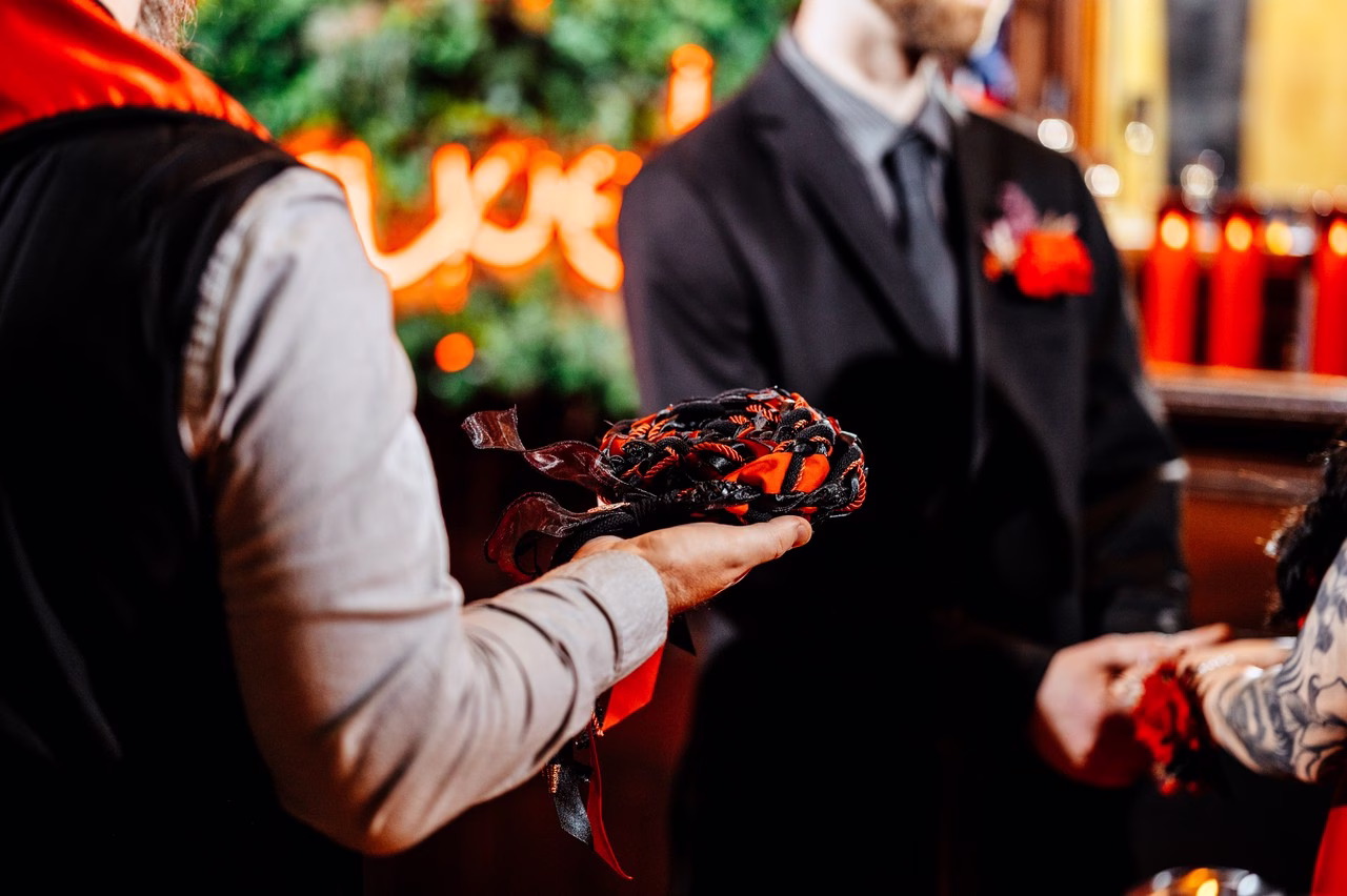 Officiant holds black and orange handfasting cords during a Satanic wedding ceremony at The Satanic Chapel, with a groom in a dark suit visible in the background
