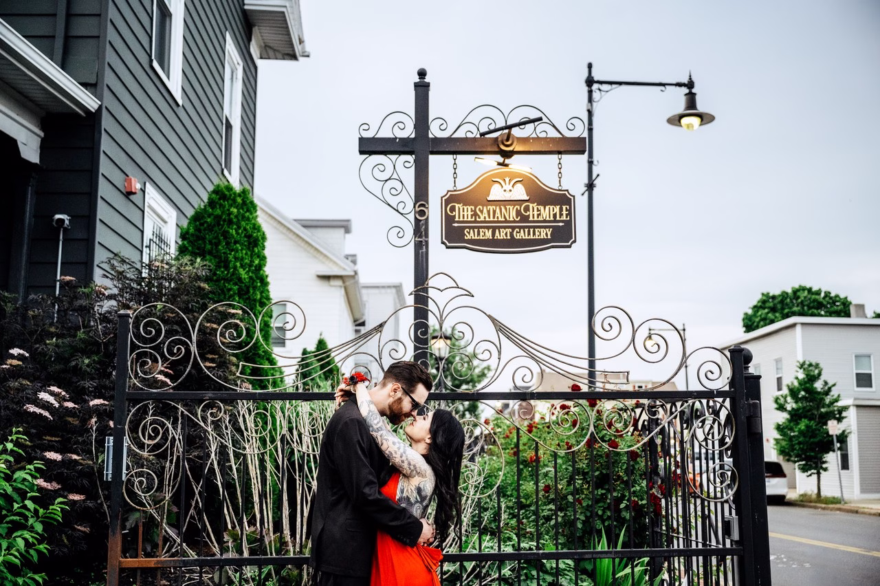 A couple sharing a kiss in front of the ornate iron gates of The Satanic Temple Salem Art Gallery, one wearing a red gown and the other in a black suit with tattoos, surrounded by red roses.