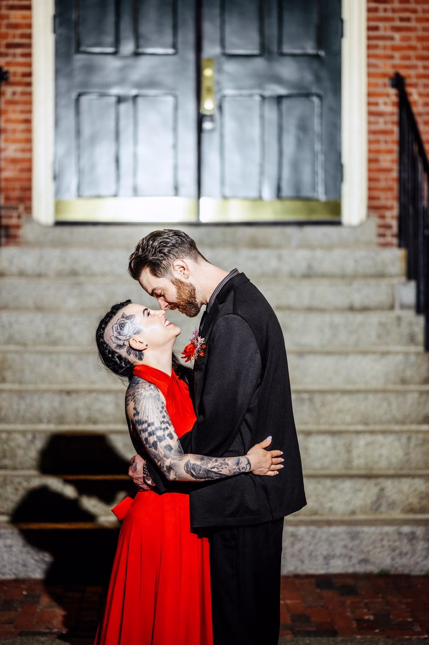A heavily tattooed bride in a red halter gown and a groom in a black suit share a romantic moment on the steps of The Satanic Chapel, photographed by Katlyn Reilly Photography