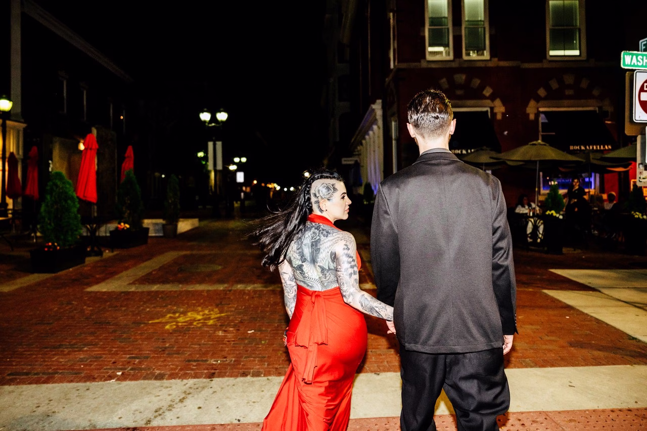 Heavily tattooed bride in backless red gown holds hands with groom in black suit while crossing a brick street at night, photographed by Katlyn Reilly Photography