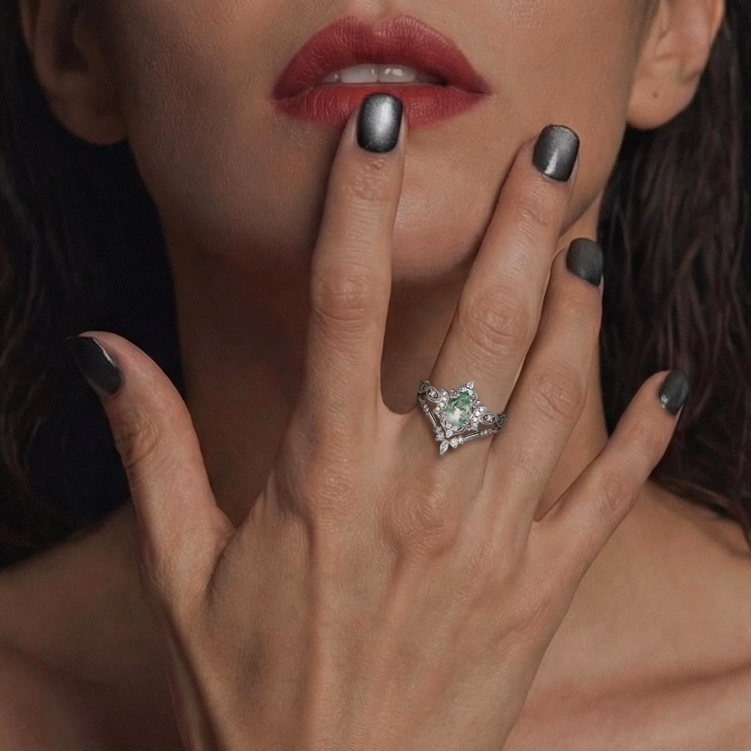 Close-up of a hand with dark metallic nail polish wearing a silver alternative engagement ring set featuring a green gemstone center stone with intricate diamond details, styled with deep red lips in background, by StarlandUS jewelry