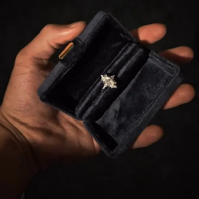 A hand holds an open black velvet ring box displaying a marquise diamond engagement ring with side stones, an alternative engagement ring by StarlandUS jewelry