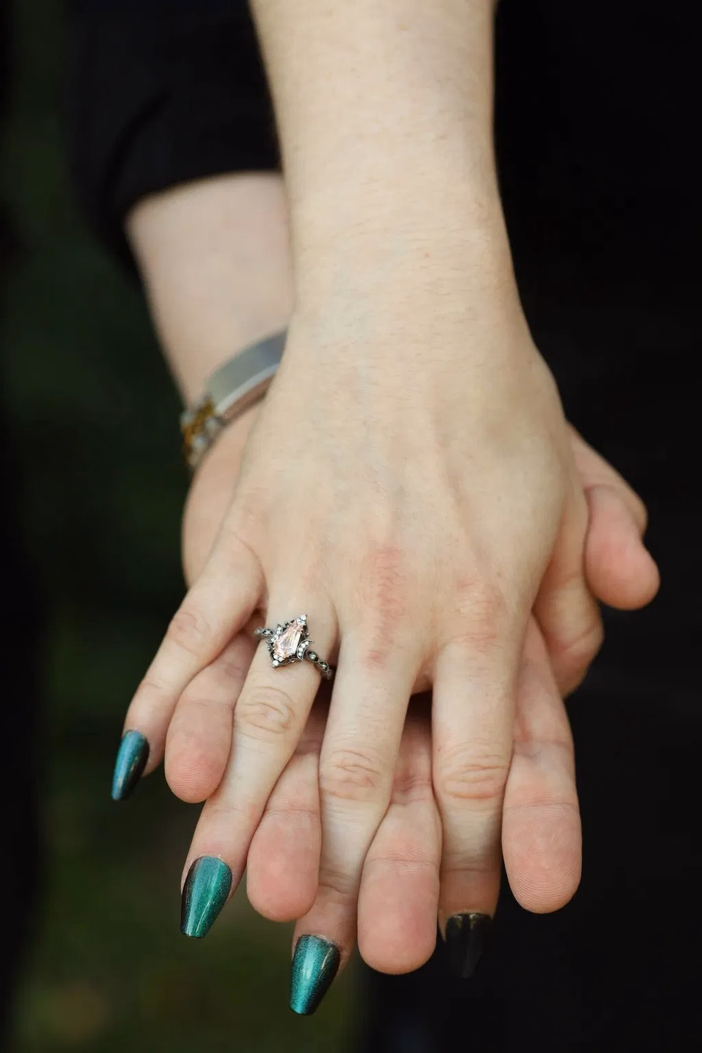 Two people holding hands showing a silver alternative engagement ring with a marquise pink stone and intricate setting, with teal chrome stiletto nails, by StarlandUS jewelry