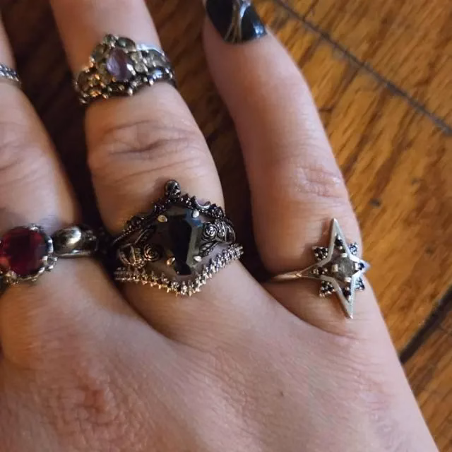 Hand wearing multiple alternative engagement rings including a gothic dark gemstone ring with ornate silver setting, a star-shaped ring, a red gemstone ring, and a crown-style ring, with dark nail polish, from StarlandUS jewelry