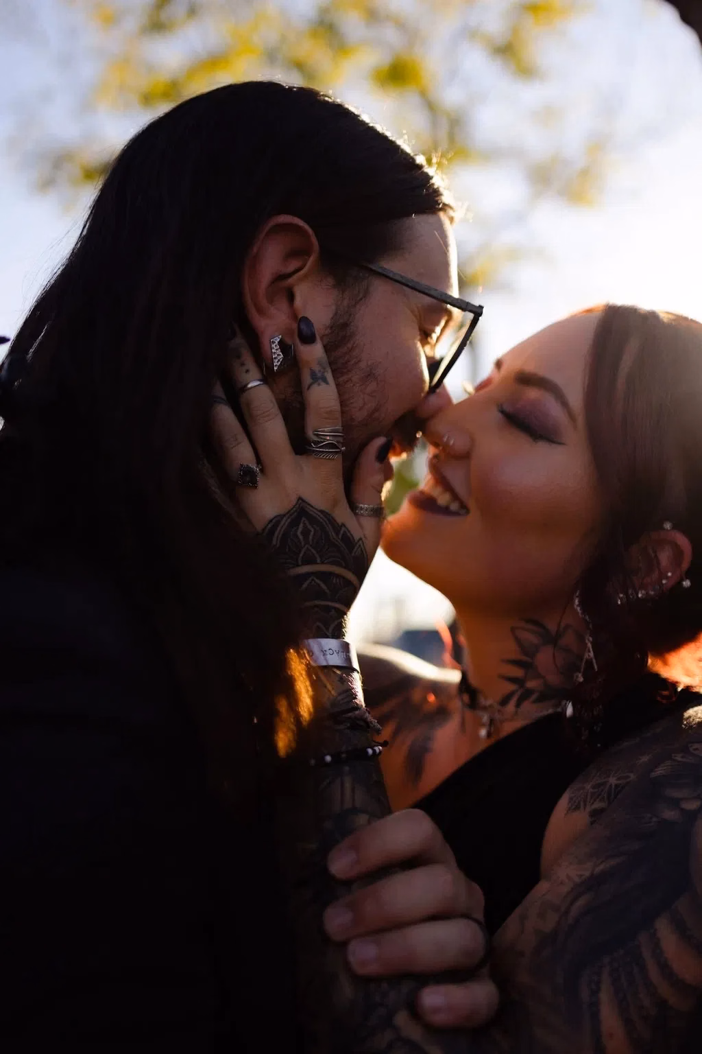 Two heavily tattooed partners share a kiss in golden hour light, with multiple alternative engagement rings and stacked silver bands visible on their hands, styled in dark alternative fashion with piercings and ear cuffs
