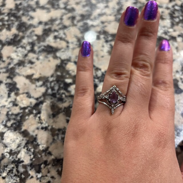 A hand with purple glitter nail polish wearing a silver ring set featuring a pear-cut amethyst center stone with diamond accents and a matching chevron band, photographed on a granite surface