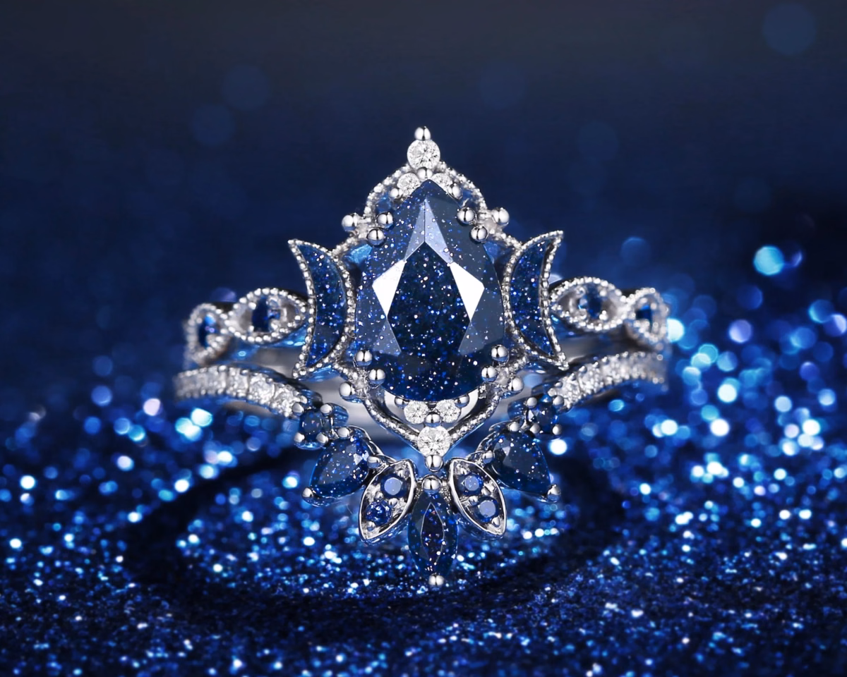 An ornate silver ring featuring a pear-cut blue sandstone center stone surrounded by blue gemstone floral and lotus motifs, small white accent stones, and milgrain detailing, styled as an alternative celestial engagement ring against a sparkling blue glitter background