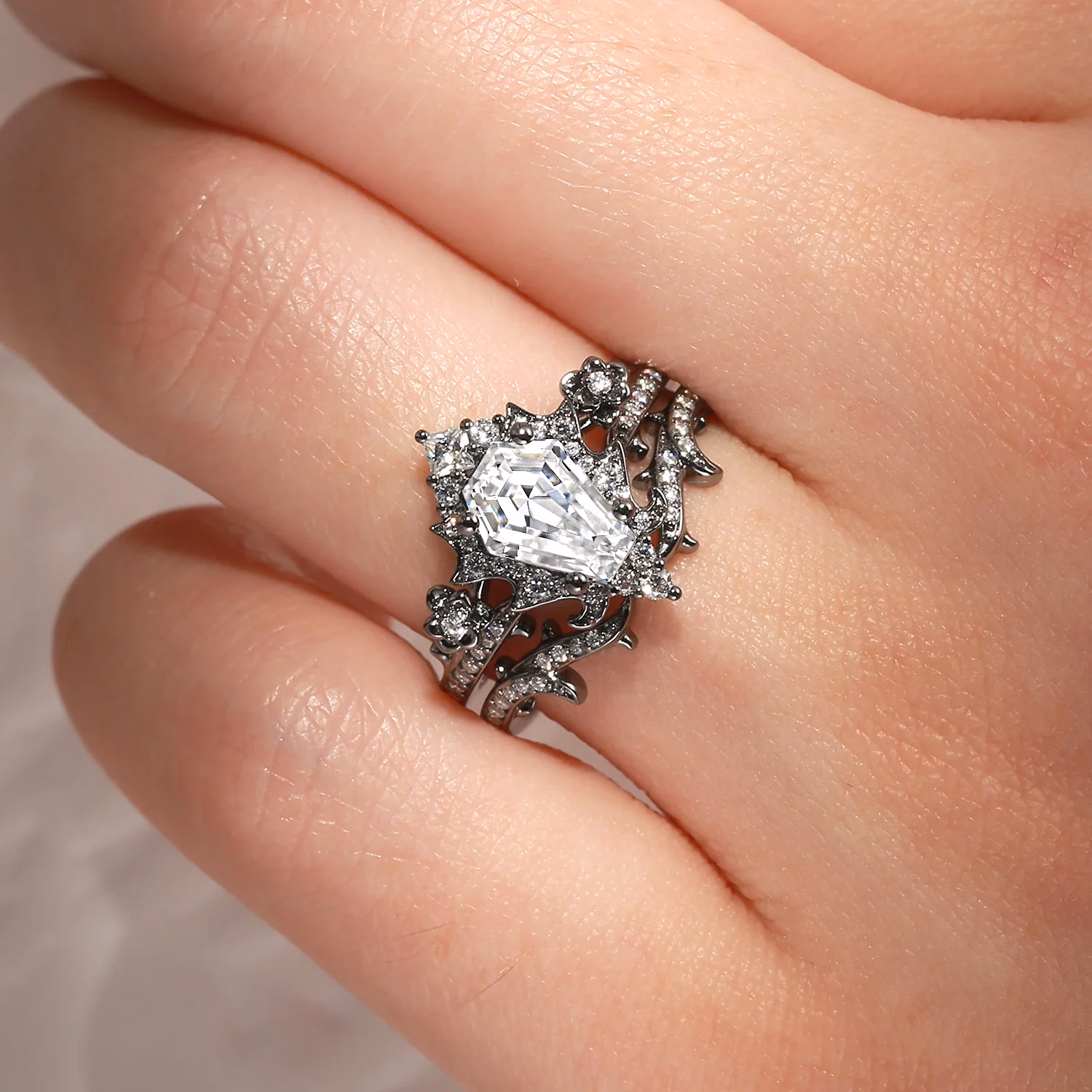 A shield-cut white moissanite center stone set in a dark oxidized black metal ring with intricate gothic floral branch detailing and pavé accent stones, worn on a hand, by StarlandUS