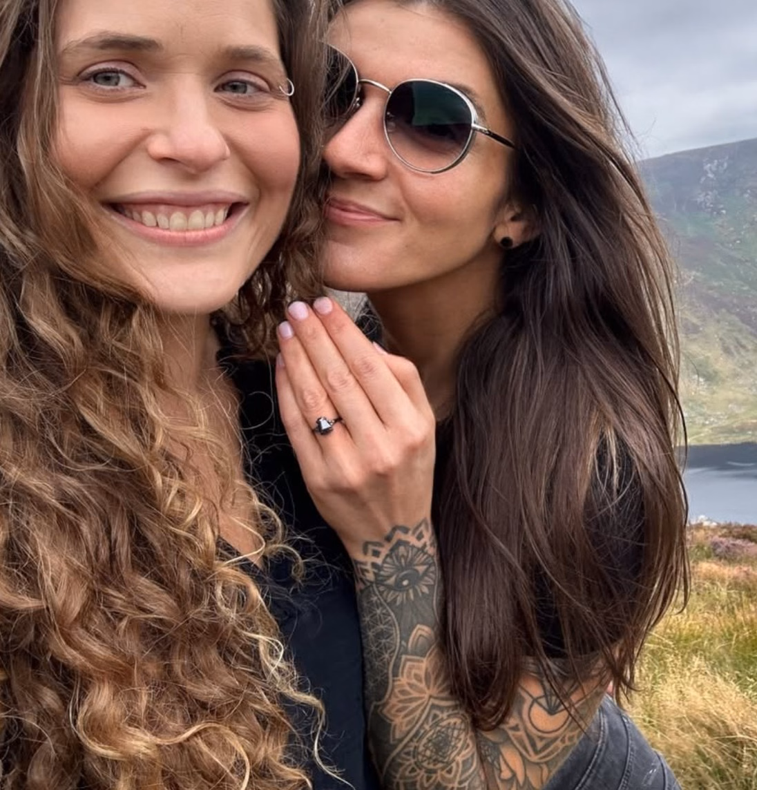 Two people posing cheek to cheek outdoors with a mountain and lake backdrop, one partner displaying an alternative engagement ring by StarlandUS on their tattooed hand