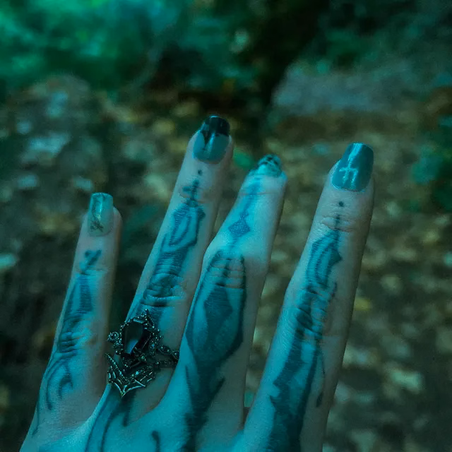 A hand with finger tattoos and teal nail polish displays an ornate gothic-style black stone ring with silver crown setting, photographed against a green forest background, ring by StarlandUS