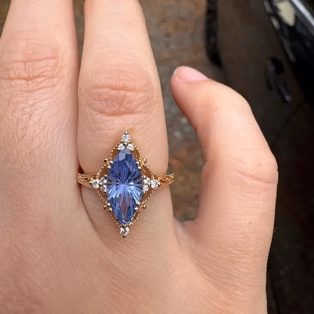 Close-up of a hand wearing an ornate rose gold ring featuring a marquise-cut blue tanzanite stone surrounded by small diamonds in a vintage-style setting, by StarlandUS