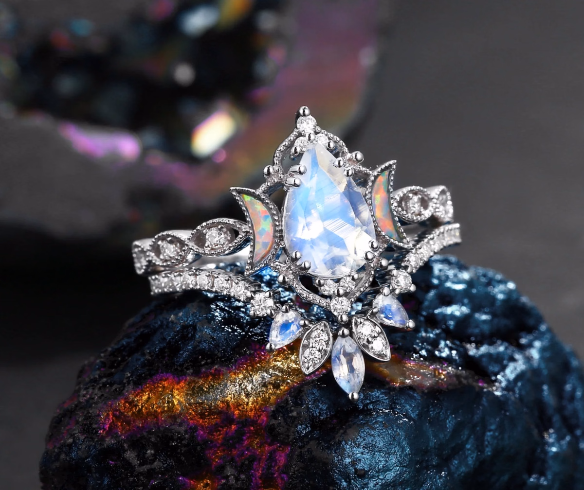 A celestial-style silver engagement ring featuring a pear-cut moonstone center stone surrounded by a halo of white sapphires, flanked by crescent moon opal side stones and marquise moonstone leaf accents, displayed on dark iridescent mineral rock