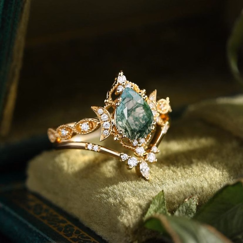 Nature-inspired yellow gold engagement ring featuring a marquise moss agate center stone surrounded by diamond halo with crescent moon and leaf details, displayed on green velvet jewelry box