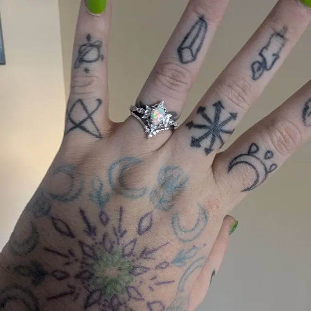 A heavily tattooed hand displaying a silver star-shaped alternative engagement ring with an opal center stone, with green nail polish visible, showcasing StarlandUS jewelry