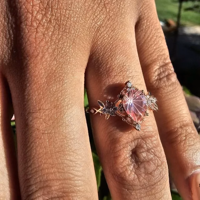 Close-up of a hand wearing a rose gold alternative engagement ring featuring a carved pink gemstone centerpiece with floral and leaf detailing and accent diamonds, by StarlandUS jewelry