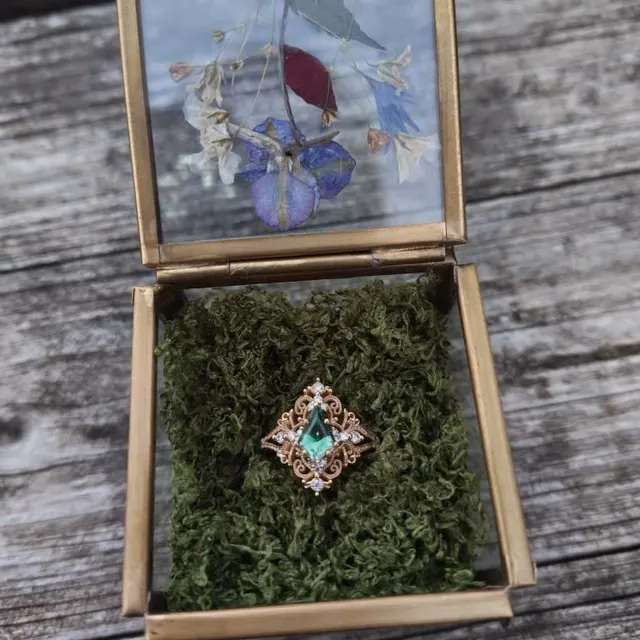 An ornate rose gold filigree ring with a pear-shaped green emerald center stone and diamond accents, displayed on green moss inside a brass and glass jewelry box with pressed flowers, an alternative engagement ring by StarlandUS